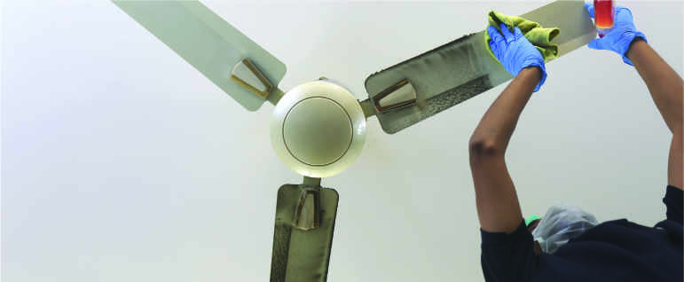 A cleaner wearing a dust mask cleaning dust off a fan using a duster and spray bottle